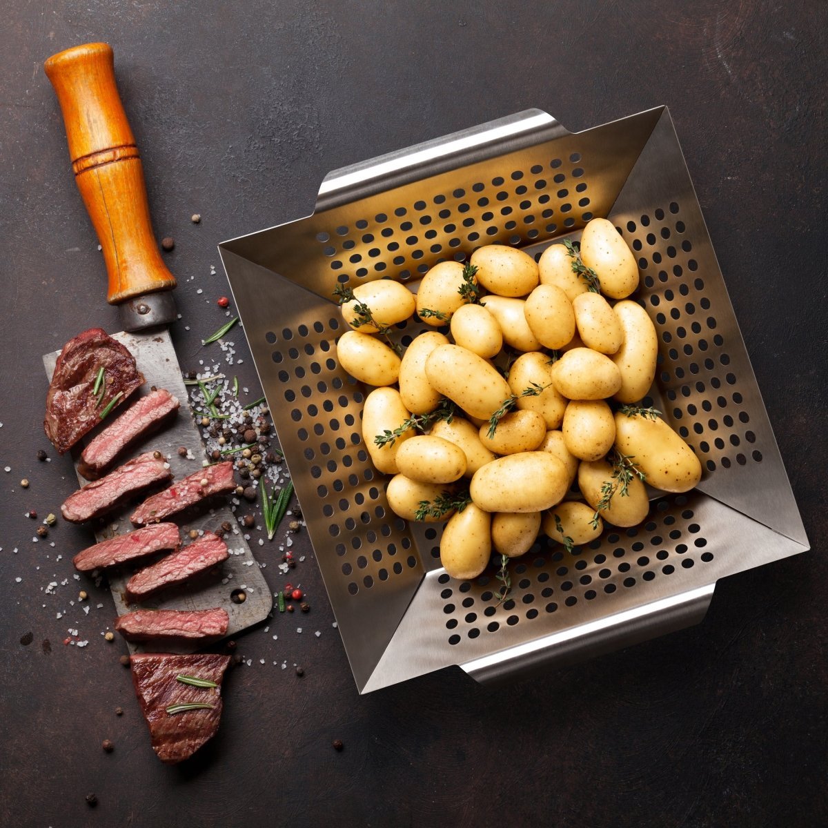 Großer Edelstahl-Grillkorb mit Kartoffeln und Steak auf dunklem Untergrund.