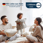 Familie mit zwei Erwachsenen und einem Kind, die auf einem Bett mit weißen Kissen spielen.