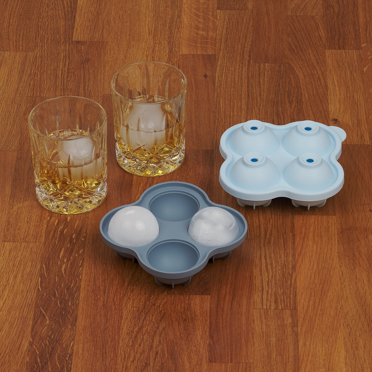 Zwei Whiskygläser mit Eiskugeln neben einer leeren und einer vollen blauen Eiskugelform auf Holzoberfläche.