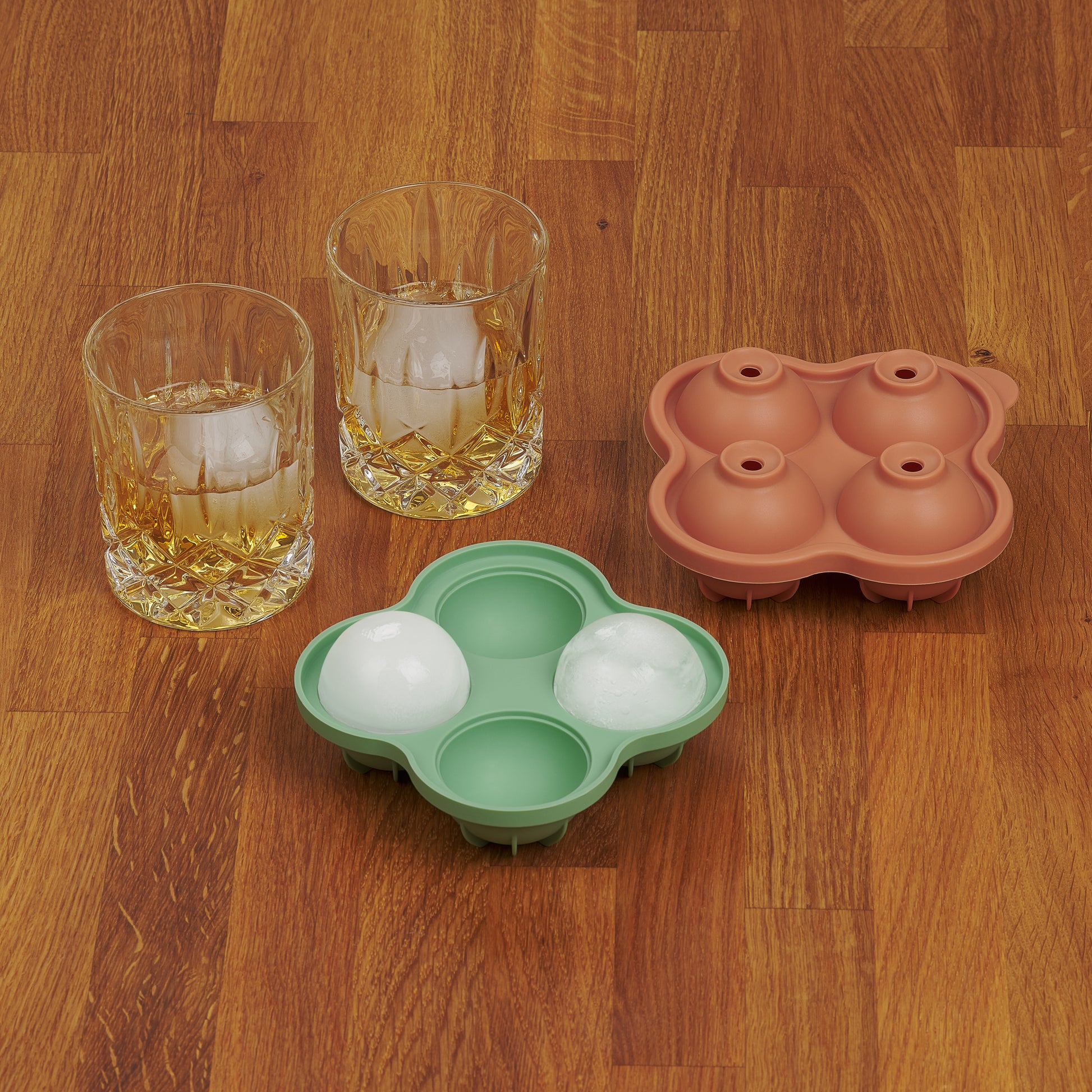 Zwei Whiskygläser mit Eiskugeln neben einer leeren und einer vollen gruenen Eiskugelform auf Holzoberfläche.