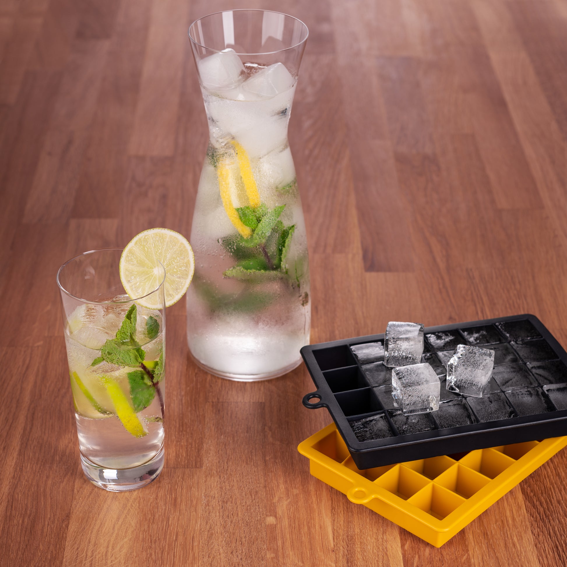 Getränke in Gläsern mit Eiswürfeln neben schwarzer und gelber Eiswürfelform auf Holztisch.