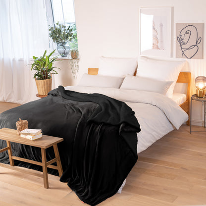 Schwarze Decke mit strukturierter Oberfläche, ausgebreitet über ein Bett in einem Raum mit natürlicher Beleuchtung, Pflanzen und dekorativen Elementen.