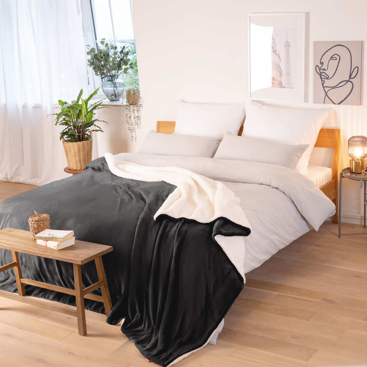 Anthrazit Decke mit weißer Fleece-Innenseite, ausgebreitet über ein Bett in einem hellen Zimmer mit Pflanzen und Bildern im Hintergrund.