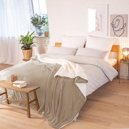Beige Decke mit weißer Fleece-Innenseite, ausgebreitet über ein Bett in einem Raum mit natürlicher Beleuchtung, Pflanzen und Bildern im Hintergrund.