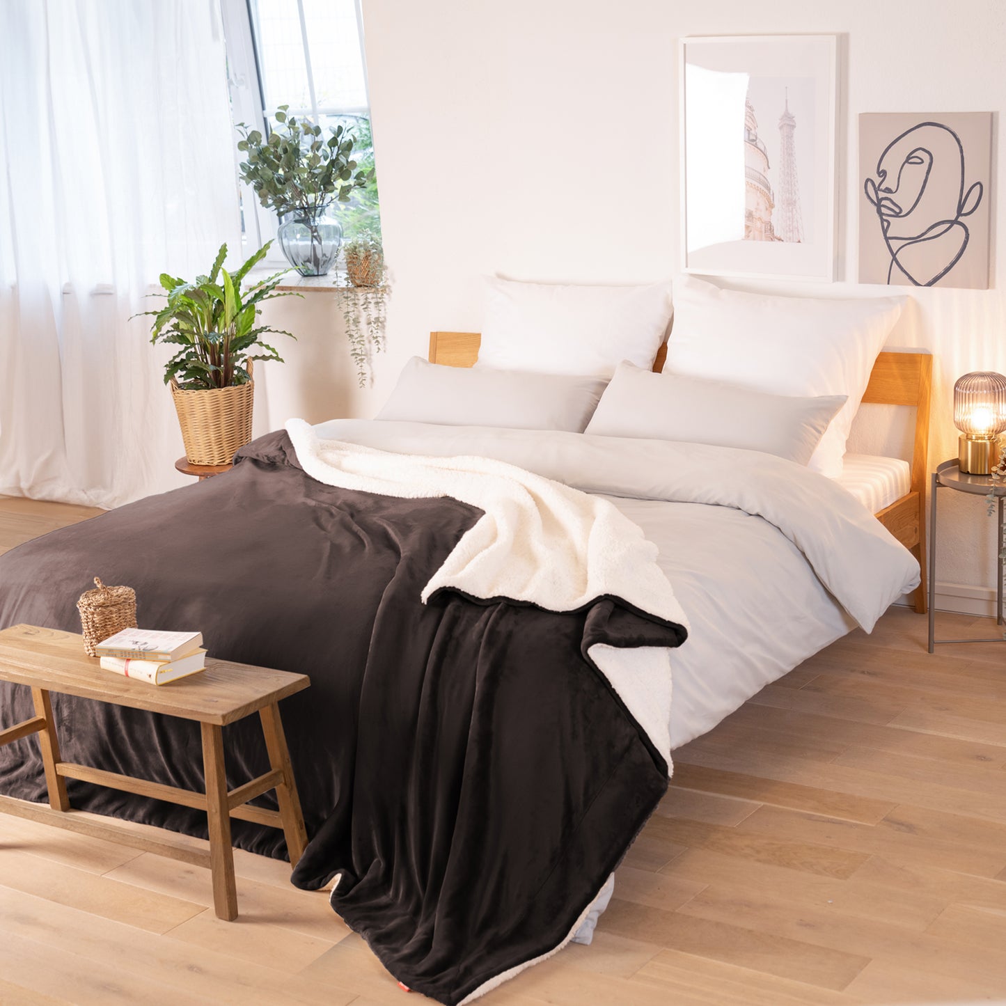 Braune Decke mit weißer Fleece-Innenseite, ausgebreitet über ein Bett in einem Raum mit natürlicher Beleuchtung, Pflanzen und Bildern im Hintergrund.