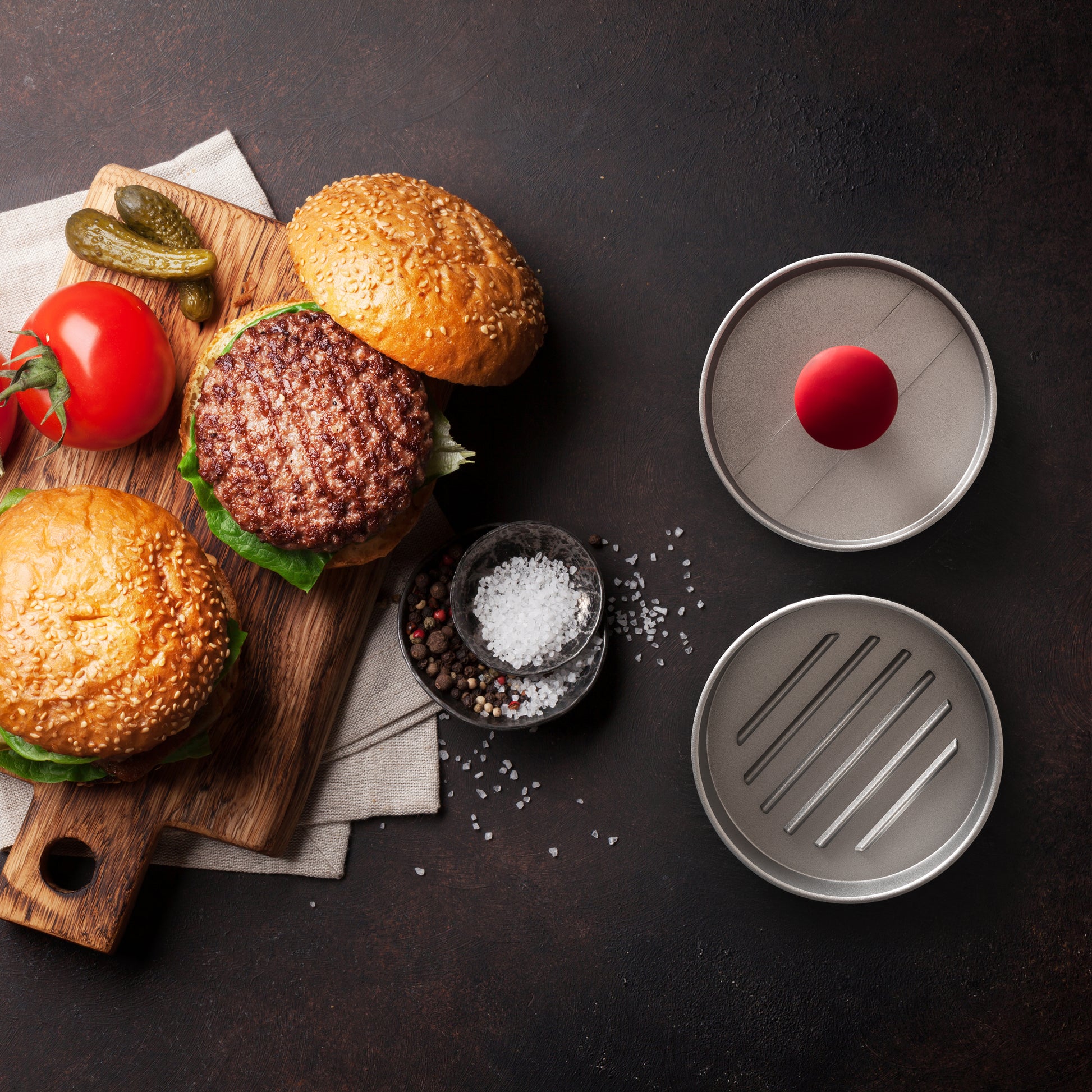 Burgerpresse mit rotem Stempel, unmontiert neben zwei fertigen Burgern auf einem Schneidebrett mit Zutaten.