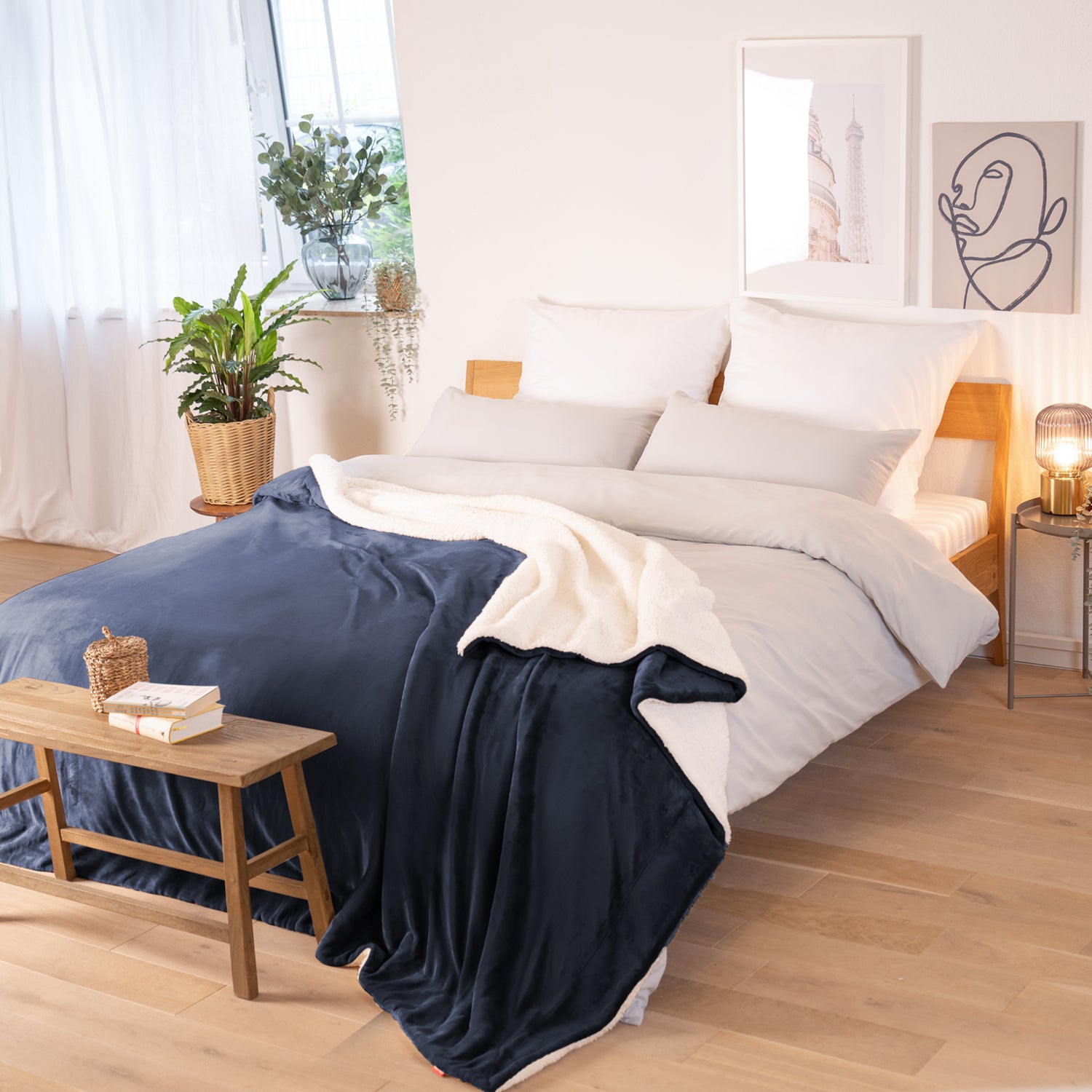Dunkelblaue Decke mit weißer Fleece-Innenseite, ausgebreitet über ein Bett in einem Raum mit natürlicher Beleuchtung, Pflanzen und Bildern im Hintergrund.