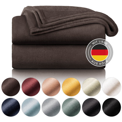 Braune Fleece-Kuscheldecke gefaltet auf einem weißen Hintergrund, umgeben von Farbmustern und mit dem Label 'Mit Liebe aus Köln'.
