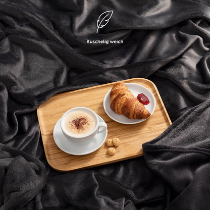 Schwarze Fleece-Kuscheldecke auf dem Bett mit einem Frühstückstablett, darauf ein Croissant und eine Tasse Kaffee, mit dem Hinweis 'Kuschelig weich'.