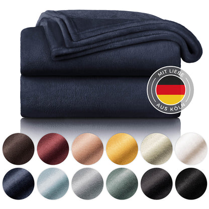 Gestapelte marineblaue Fleece-Kuscheldecken mit dem Label 'Mit Liebe aus Köln', umgeben von Farbmustern in verschiedenen Tönen wie Dunkelbraun, Weinrot, Beige, Gelb, Olivgrün, Grau und Schwarz.