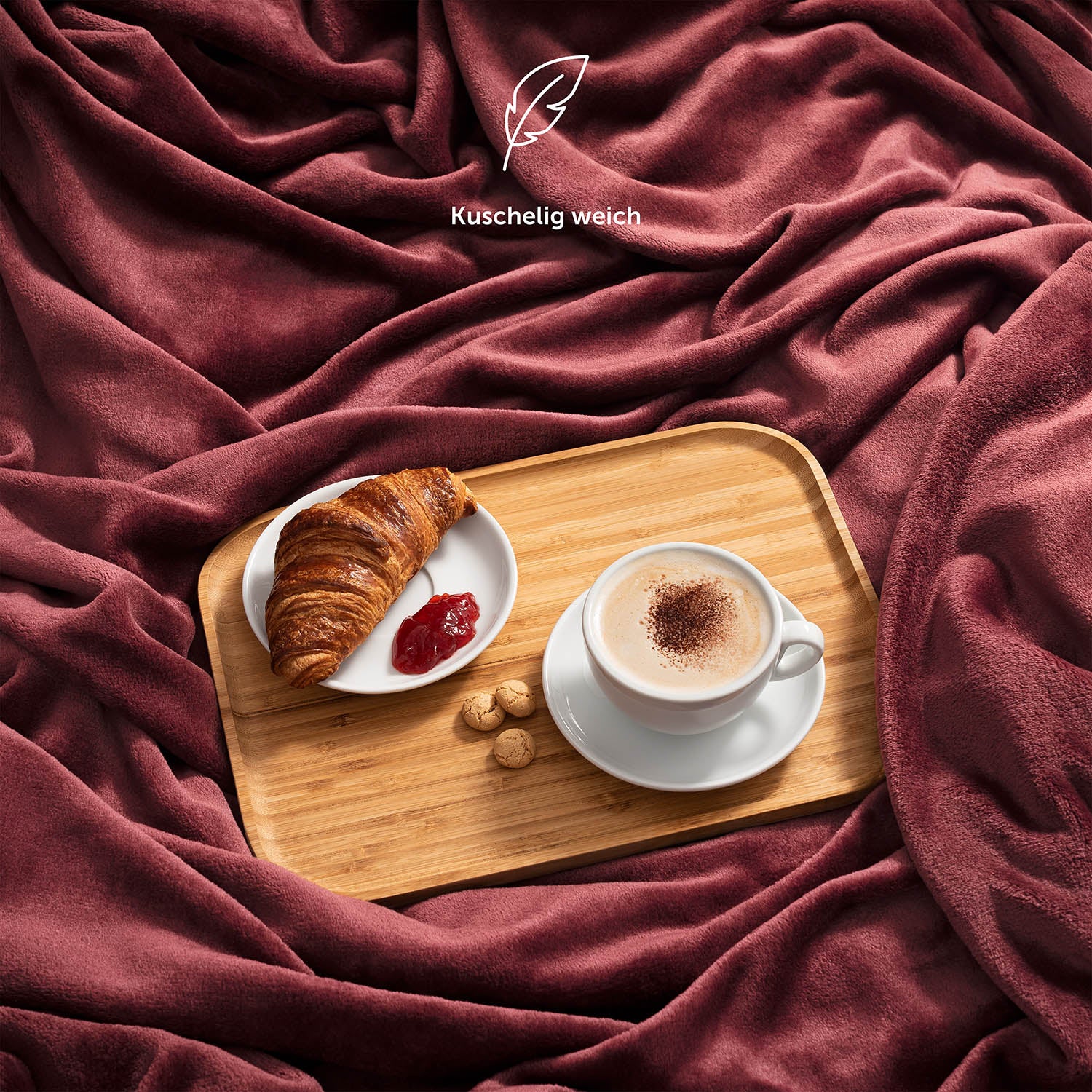 Weinrote Fleece-Kuscheldecke auf einem Bett mit einem Serviertablett mit Frühstück, bestehend aus Kaffee und Croissant, und der Beschreibung 'Kuschelig weich'.