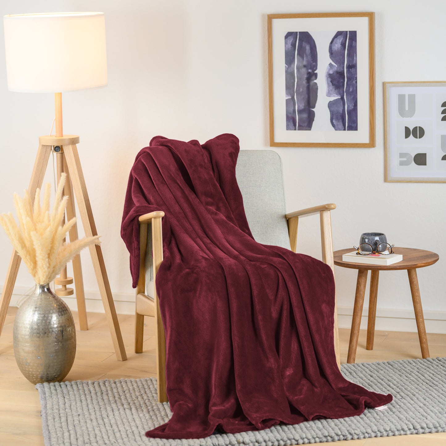 Weinrote Fleece-Kuscheldecke stilvoll über einen Sessel in einem Wohnzimmer drapiert, neben einem Beistelltisch mit einer Vase und einer Brille.