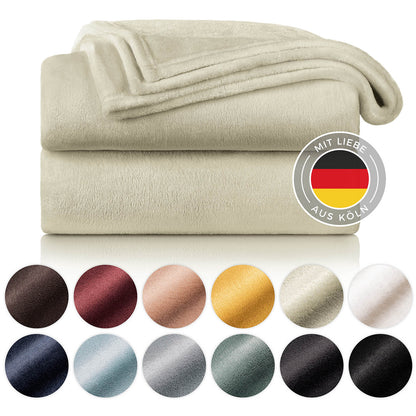 Gestapelte cremefarbene Fleece-Kuscheldecken mit Farbmustern und dem Siegel 'Mit Liebe aus Köln'.