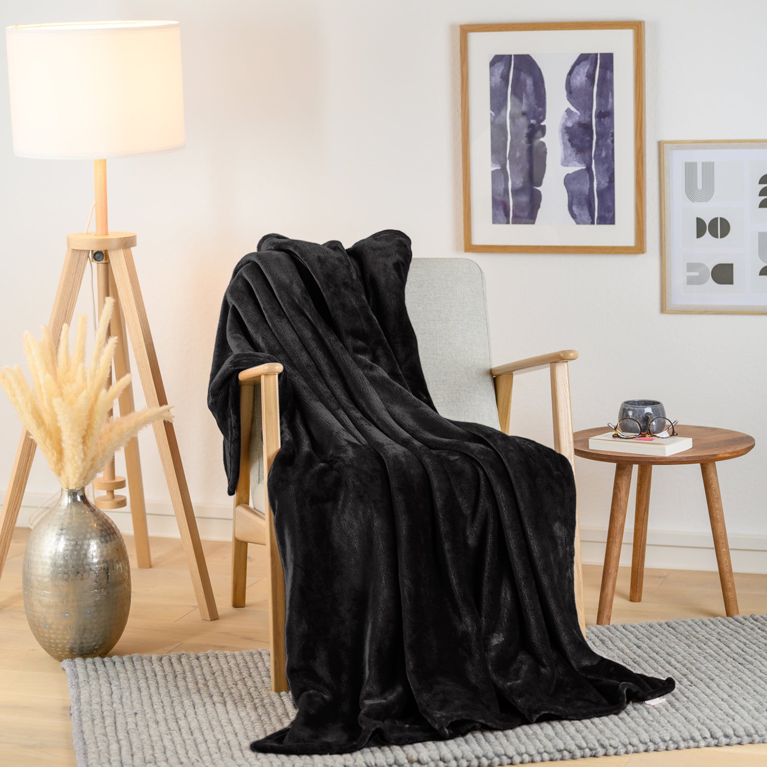 Gemütlich eingerichtetes Wohnzimmer mit einer schwarzen Fleece-Kuscheldecke auf einem hellen Sessel. Neben dem Sessel steht eine Holzlampe und ein kleiner Beistelltisch mit einer Vase und einer Brille darauf.