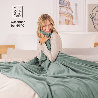 Glückliche Frau, eingehüllt in eine salbeigrüne Fleece-Kuscheldecke, sitzt auf einem Bett, mit einem Piktogramm für Waschbarkeit bei 40 °C.