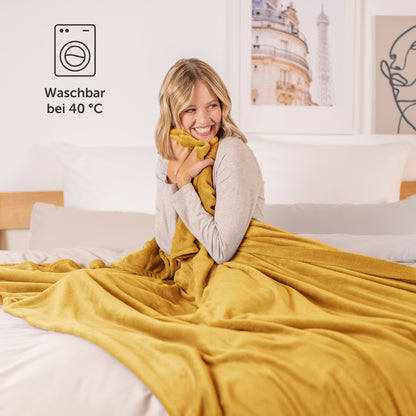 Lächelnde Frau umhüllt mit einer senfgelben Fleece-Kuscheldecke im Bett, mit einem Waschsymbol und der Temperaturangabe 40°C.