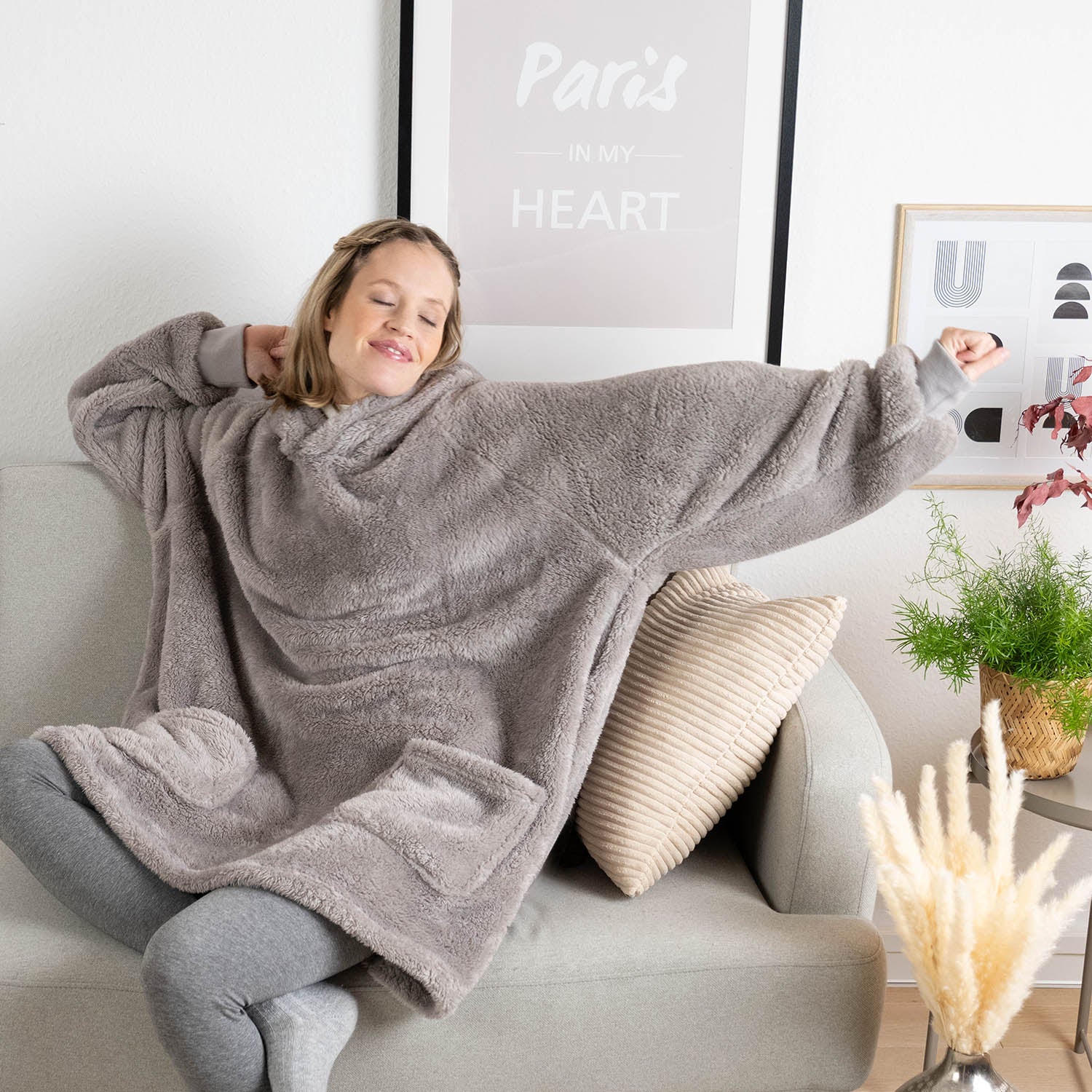 Frau sitzt entspannt auf einem Sofa, eingehüllt in eine graue Flausch-Kuscheljacke, mit Kissen, einem Buch und Pflanzen um sie herum.