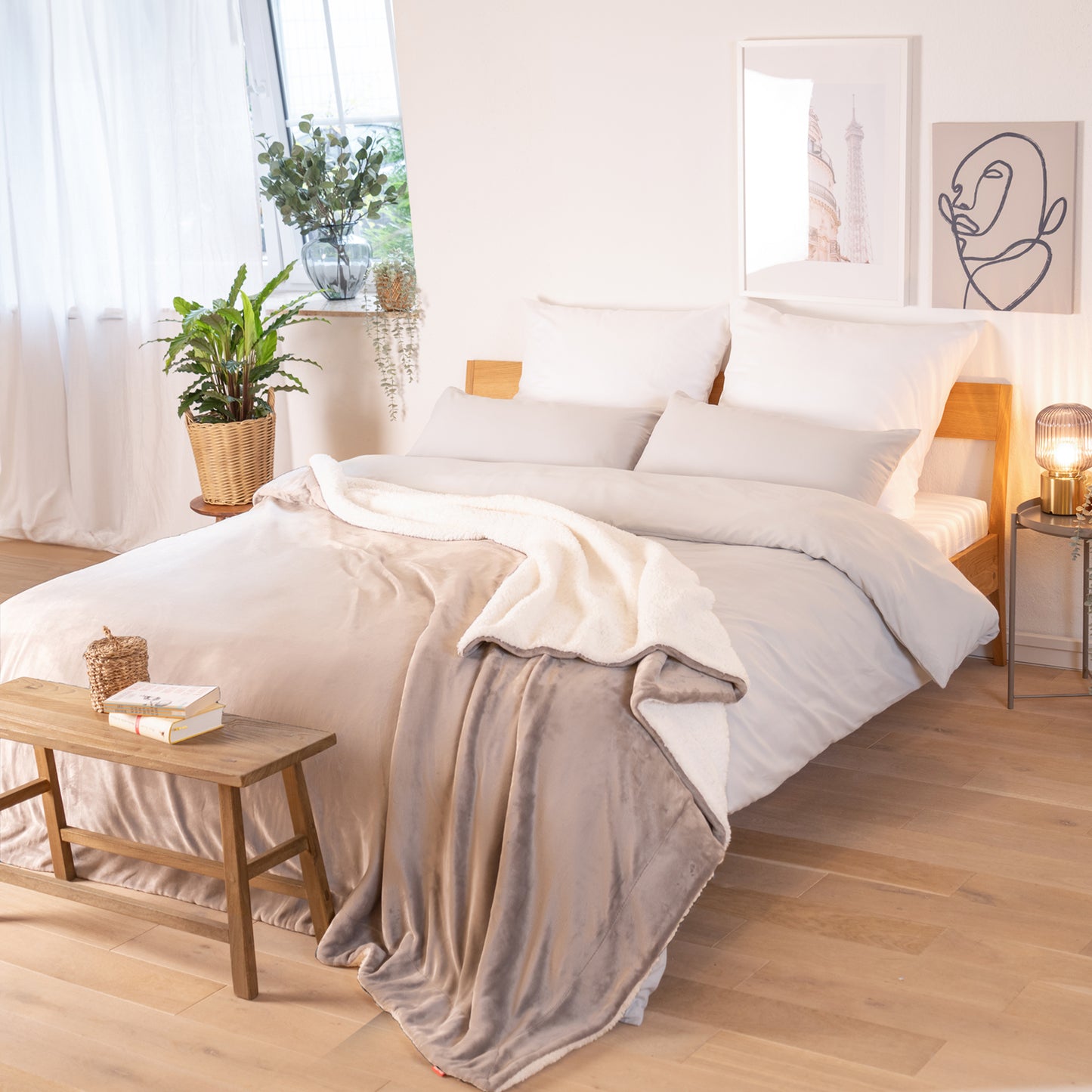 Graue Decke mit weißer Fleece-Innenseite, ausgebreitet über ein Bett in einem Raum mit natürlicher Beleuchtung, Pflanzen und Bildern im Hintergrund.