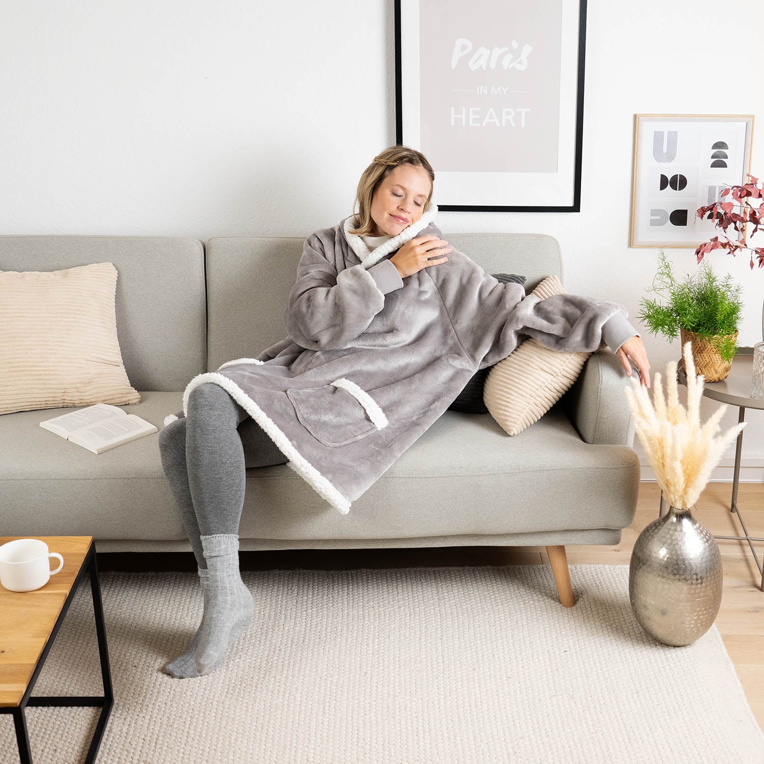 Frau sitzt entspannt auf einem Sofa, eingehüllt in eine graue Sherpa-Kuscheljacke, mit Kissen, einem Buch und Pflanzen um sie herum, im Hintergrund ein Bild mit der Aufschrift &quot;Paris in my Heart&quot;.