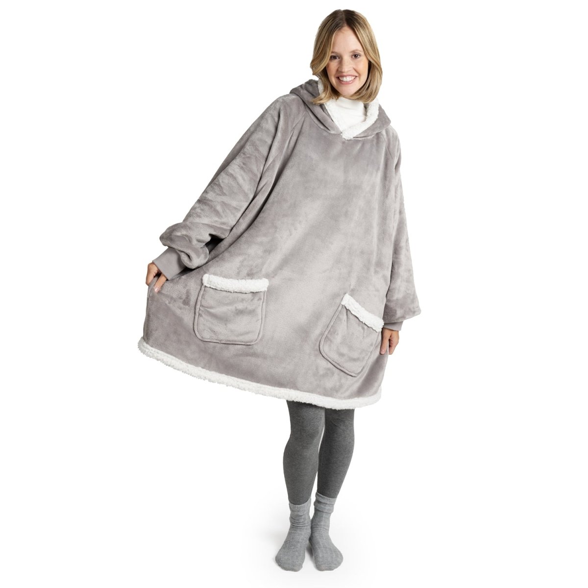 Frau in grauer Leggings und Socken lächelt, während sie eine graue Sherpa-Kuscheljacke mit weißen Saumdetails und großen Taschen trägt.