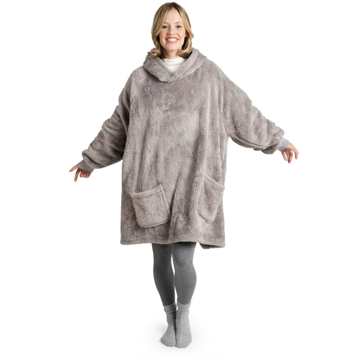 Frau in grauer Leggings und Socken lächelt, während sie eine graue Flausch-Kuscheljacke mit Kapuze und großen Taschen trägt.