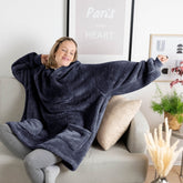 Frau entspannt auf einem Sofa, trägt eine blaue Kuscheljacke und streckt die Arme, neben ihr Kissen und eine Pflanze, im Hintergrund ein Bild mit dem Text "Paris in my Heart".