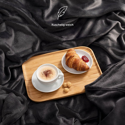 Eine marineblaue Fleece-Kuscheldecke auf einem Bett, mit einem Serviertablett, das ein Frühstück mit Kaffee und Croissant präsentiert, und der Beschreibung &