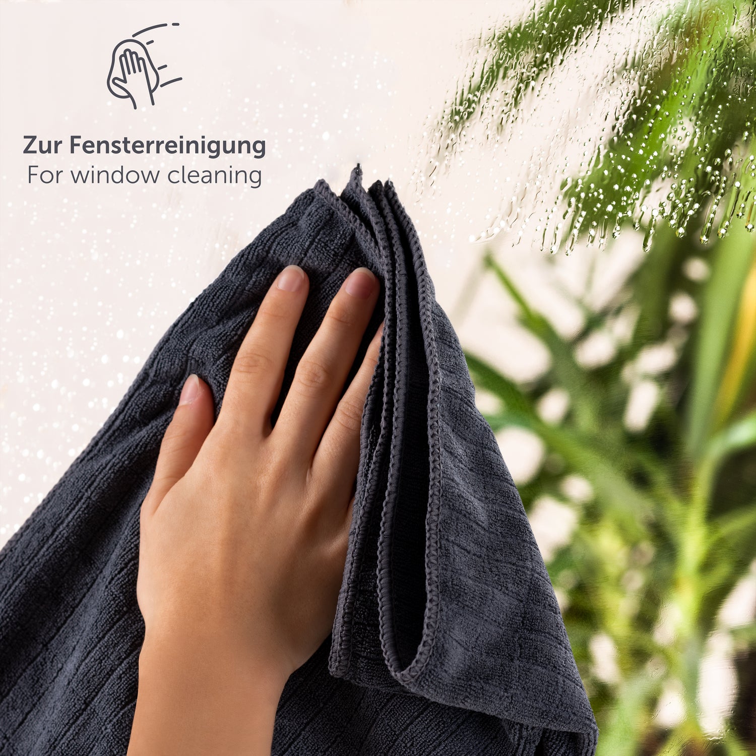 Hand hält ein Mikrofasertuch zur Fensterreinigung gegen eine Fensterscheibe mit Wassertropfen.