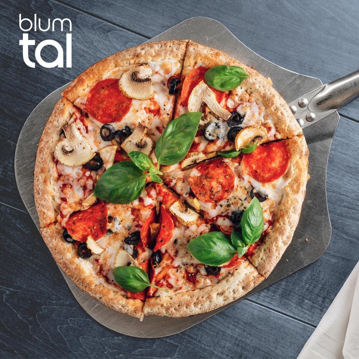 Frisch gebackene Pizza mit Salami, Pilzen, Oliven und Basilikumblättern auf einer Aluminiumschaufel von Blumtal.
