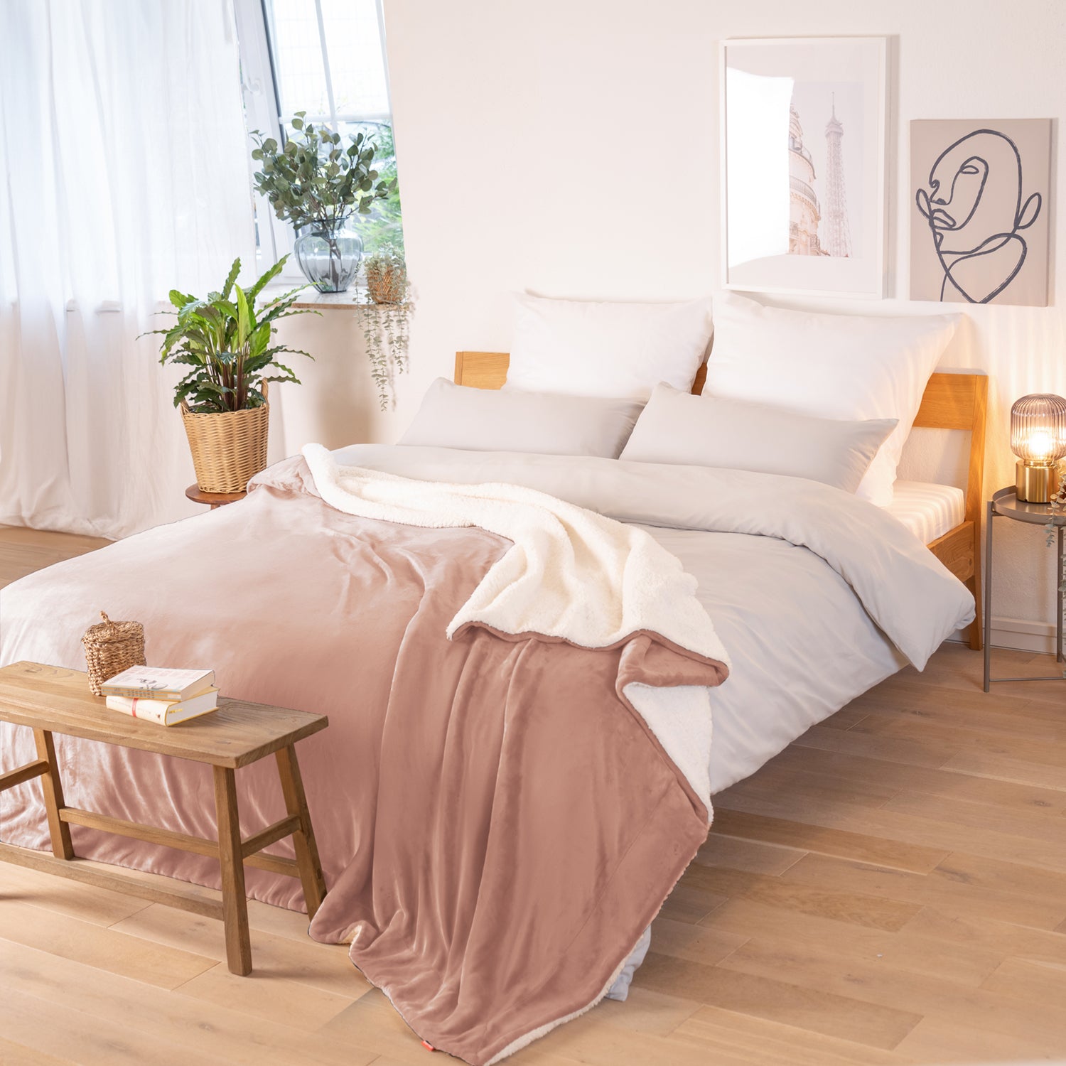 Rosa Decke mit weißer Fleece-Innenseite, ausgebreitet über ein Bett in einem Raum mit natürlicher Beleuchtung, Pflanzen und Bildern im Hintergrund.