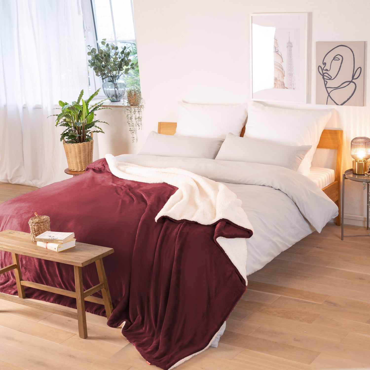 Rot-weinrote Decke mit weißer Fleece-Innenseite, ausgebreitet über ein Bett in einem Raum mit natürlicher Beleuchtung, Pflanzen und Bildern im Hintergrund.