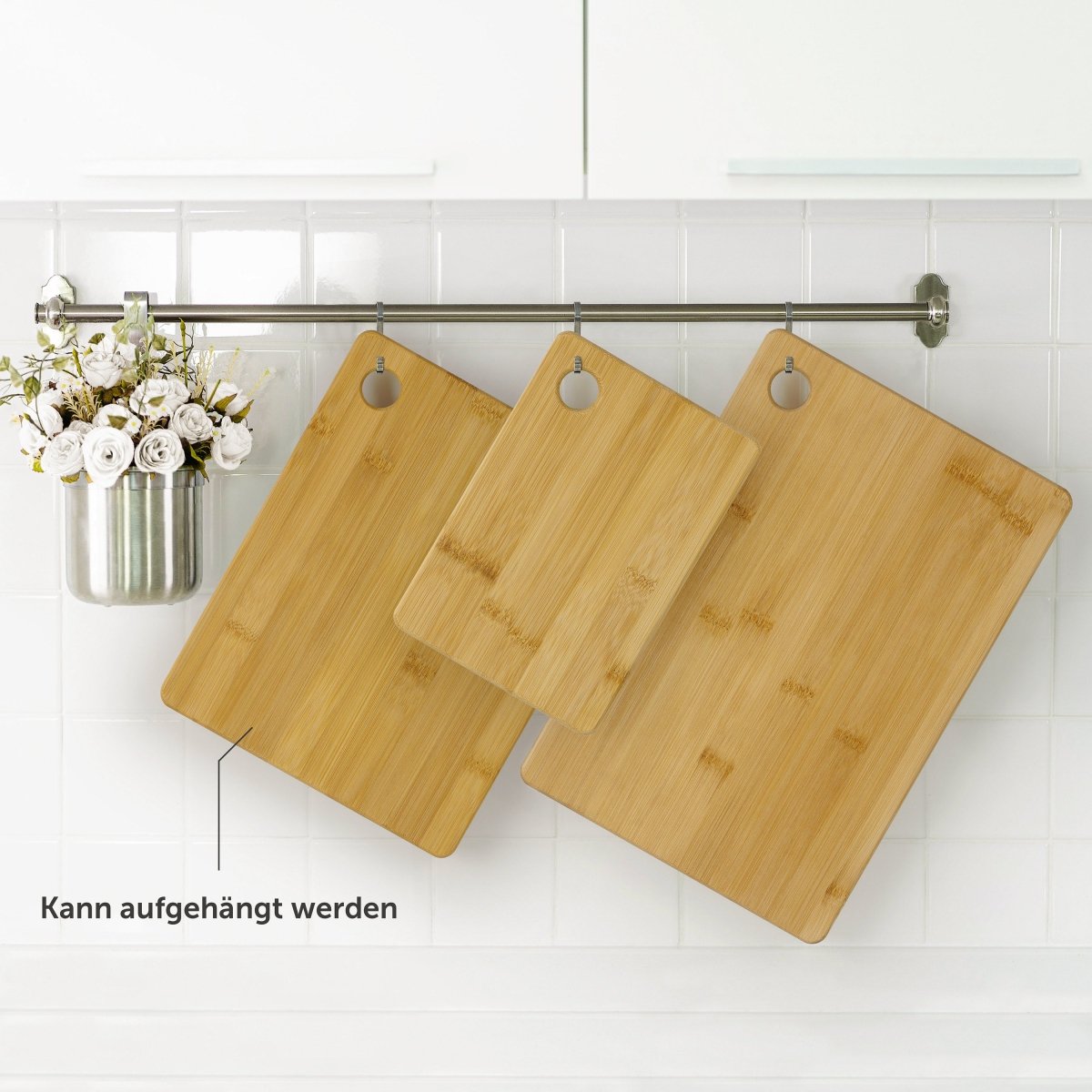 Drei Bambusschneidebretter hängen an einer Küchenleiste, neben einem Blumenarrangement.