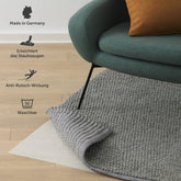 Eine Antirutschmatte unter einem grauen Teppich auf Holzboden, "Made in Germany", waschbar bei 30 Grad und mit Anti-Rutsch-Wirkung, die das Staubsaugen erleichtert.