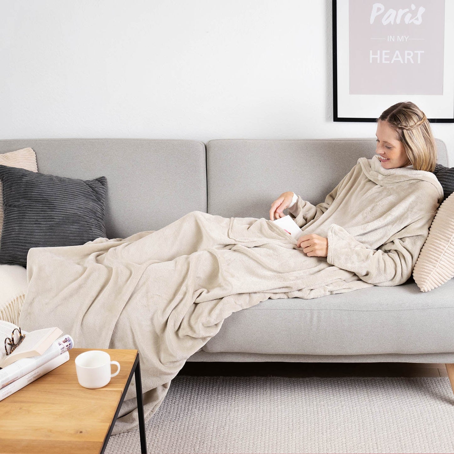 Frau liegt auf einem grauen Sofa eingehüllt in ein beige flauschiges Kleidungsstück und liest ein Buch mit einer Tasse auf einem Holztisch daneben.