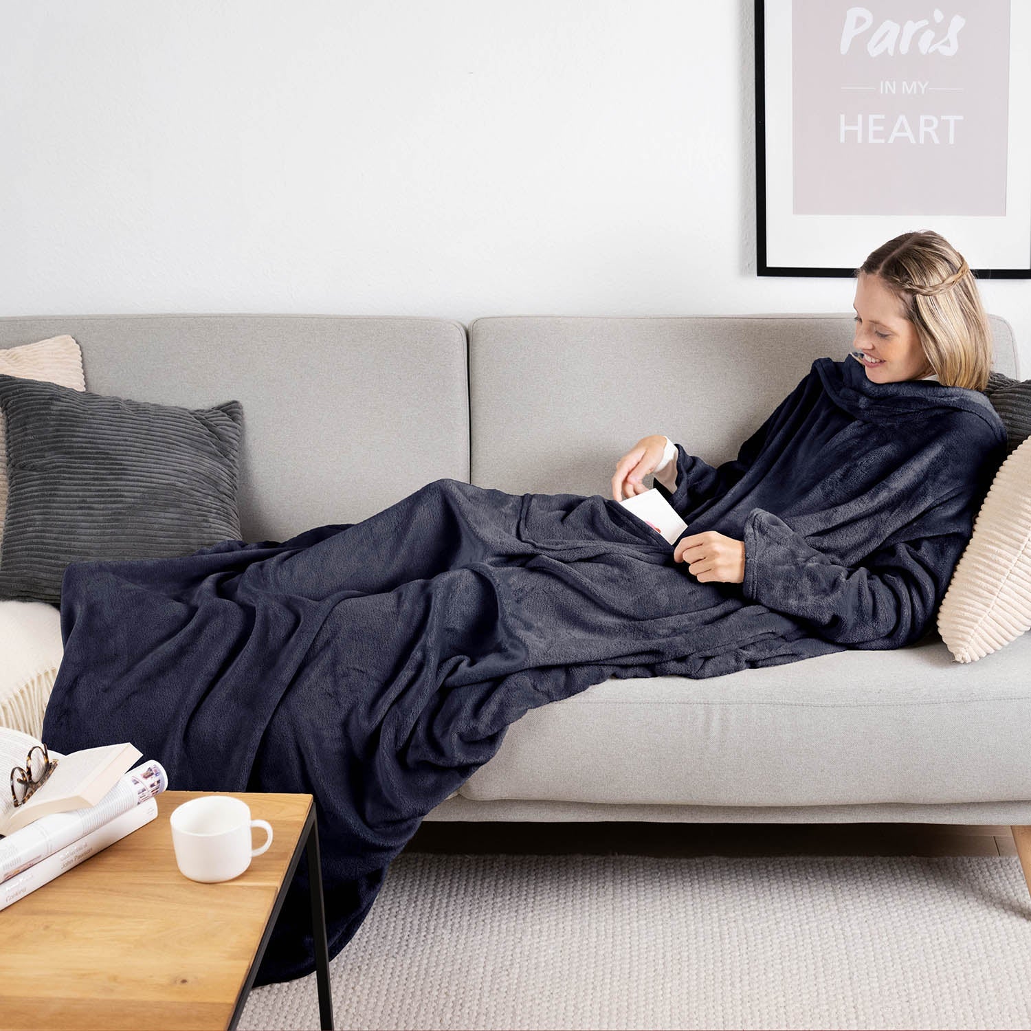 Frau liegt auf einem grauen Sofa eingehüllt in ein dunkelblau flauschiges Kleidungsstück und liest ein Buch mit einer Tasse auf einem Holztisch daneben.