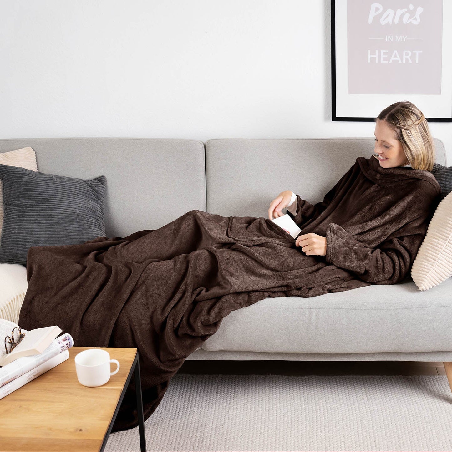 Frau liegt auf einem grauen Sofa eingehüllt in ein dunkelbraunen flauschiges Kleidungsstück und liest ein Buch mit einer Tasse auf einem Holztisch daneben.