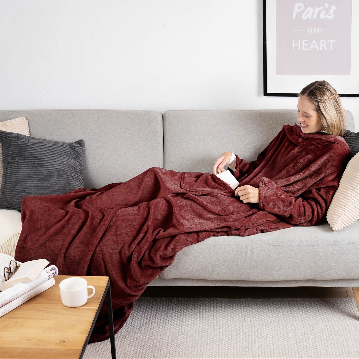 Frau liegt auf einem grauen Sofa eingehüllt in ein dunkelroten flauschiges Kleidungsstück und liest ein Buch mit einer Tasse auf einem Holztisch daneben.