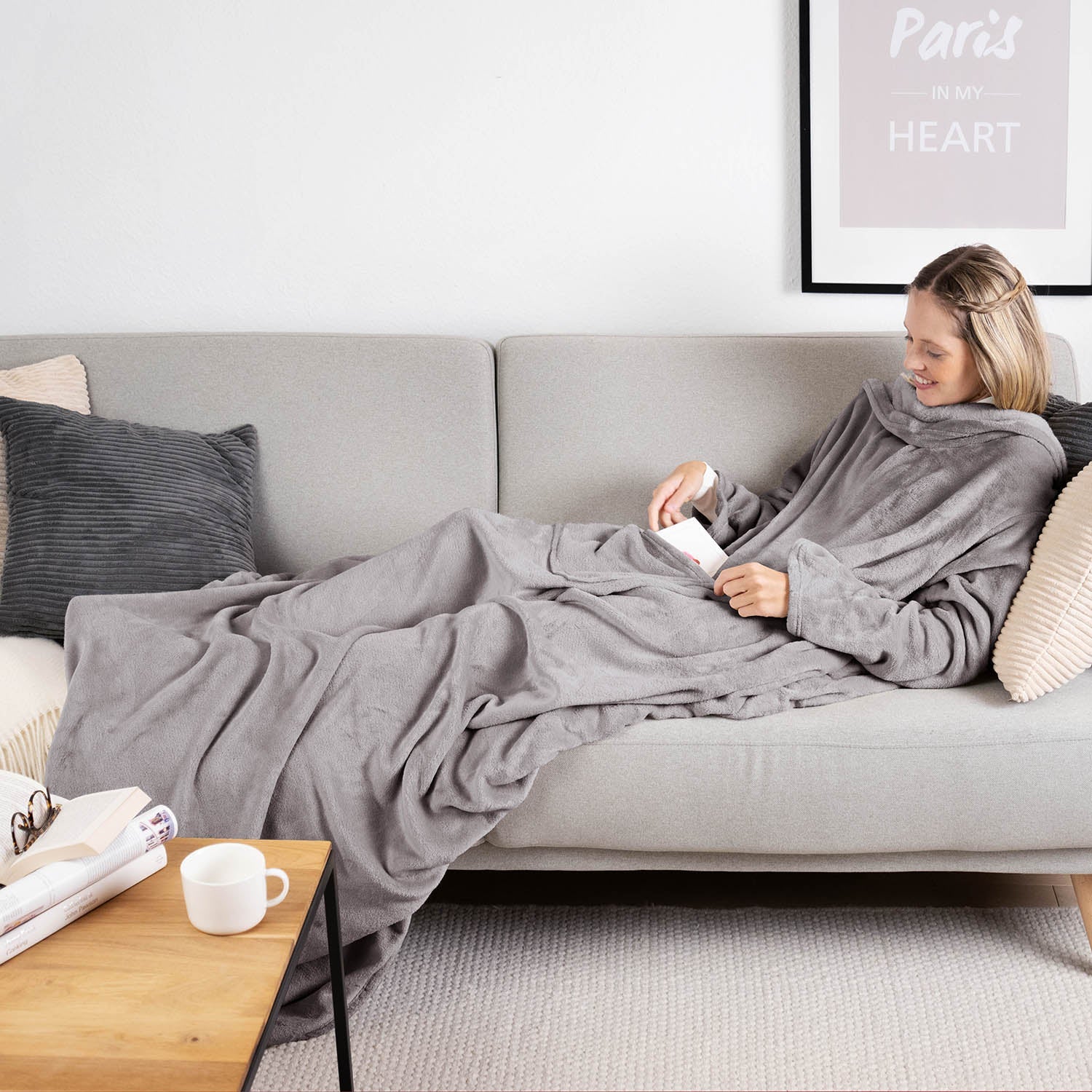 Frau liegt auf einem grauen Sofa eingehüllt in ein grauen flauschiges Kleidungsstück und liest ein Buch mit einer Tasse auf einem Holztisch daneben.