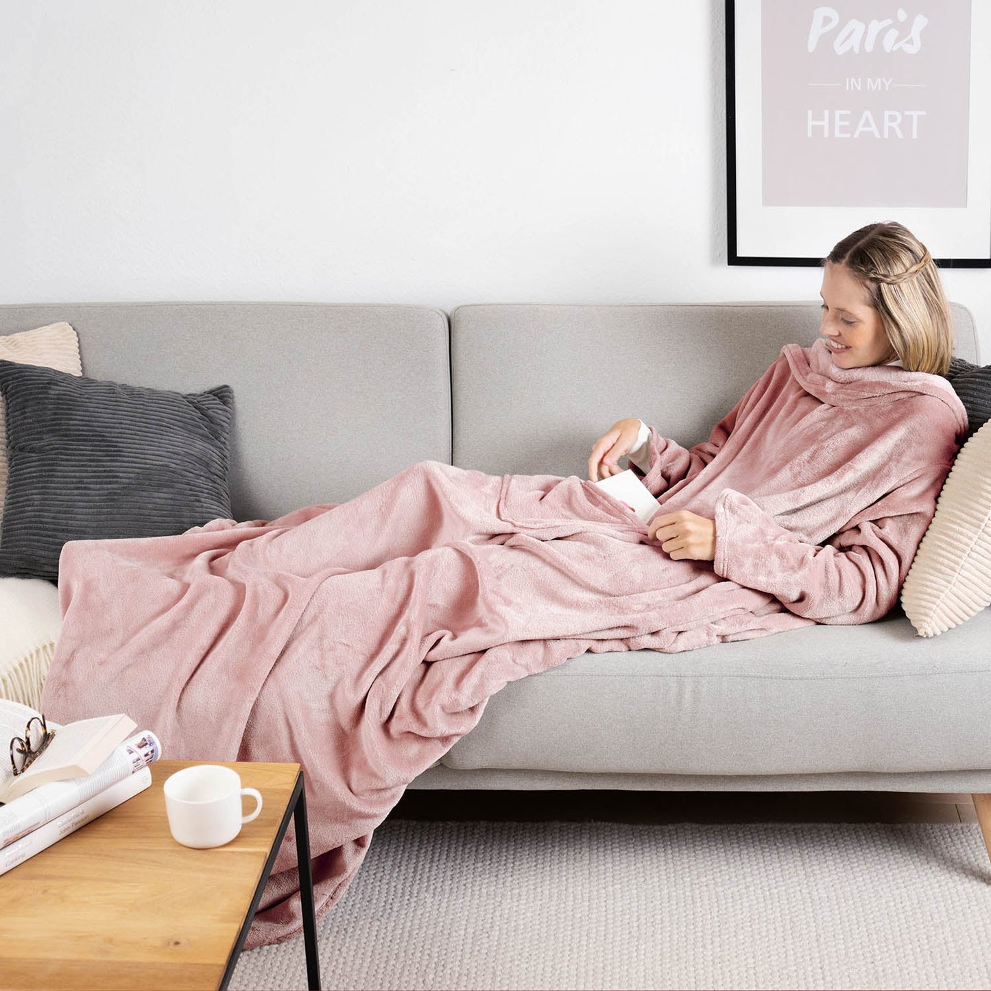 Frau liegt auf einem grauen Sofa eingehüllt in ein rosa flauschiges Kleidungsstück und liest ein Buch mit einer Tasse auf einem Holztisch daneben.
