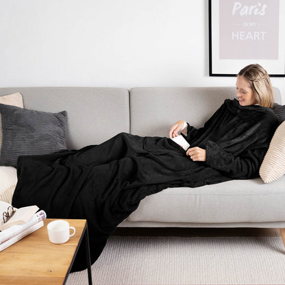 Frau liegt auf einem grauen Sofa eingehüllt in ein schwarz flauschiges Kleidungsstück und liest ein Buch mit einer Tasse auf einem Holztisch daneben.